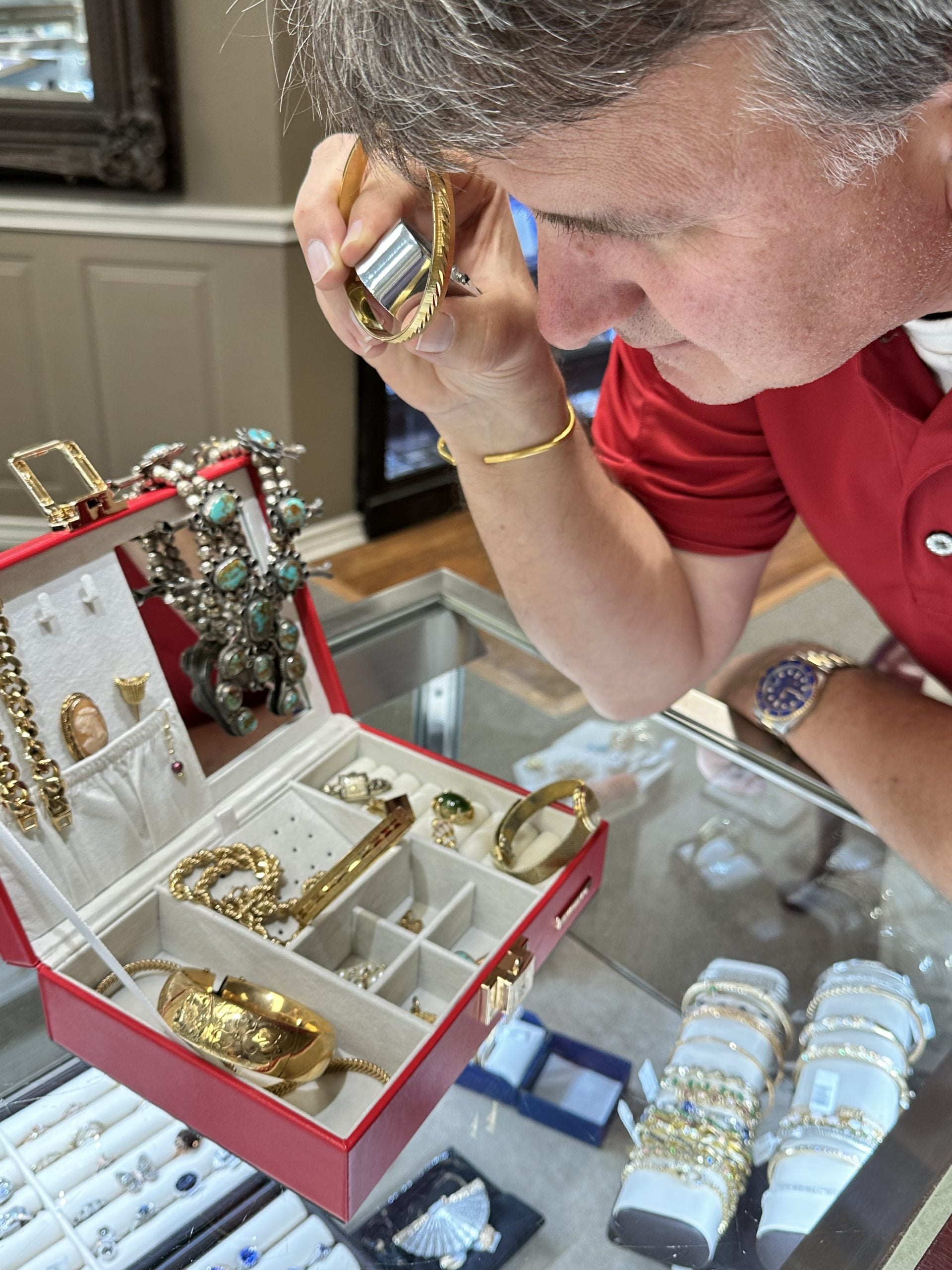 A man inspects jewelry with a loupe beside a jewelry display counter.