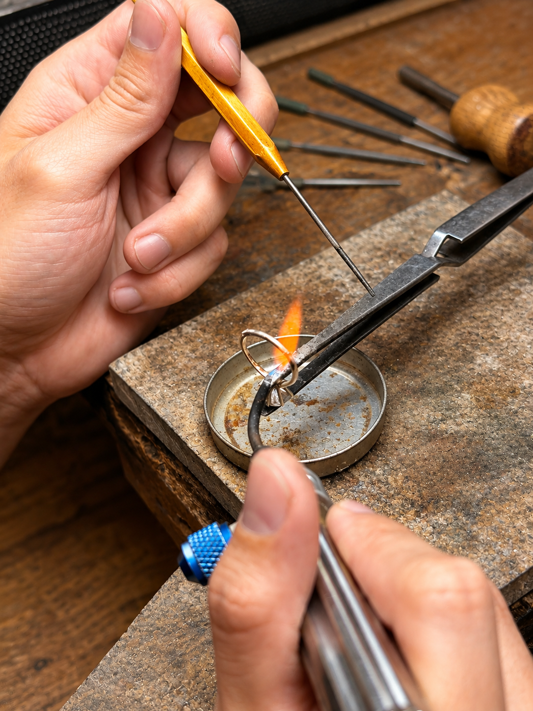 A jeweler uses a torch and hand tools to work on a ring at a repair bench.
